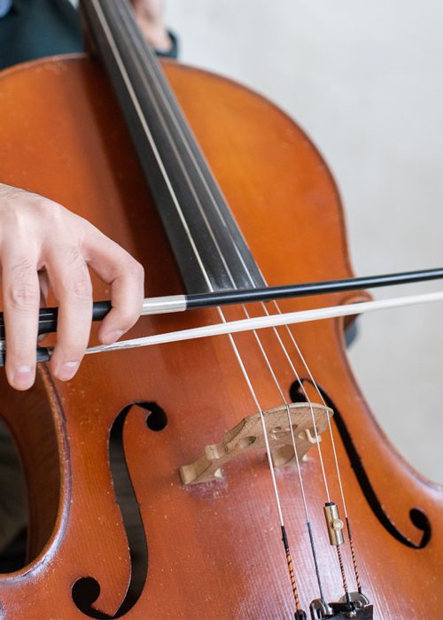 Hand of young cello player gliding fiddlestick across strings while performing piece of music in front of camera